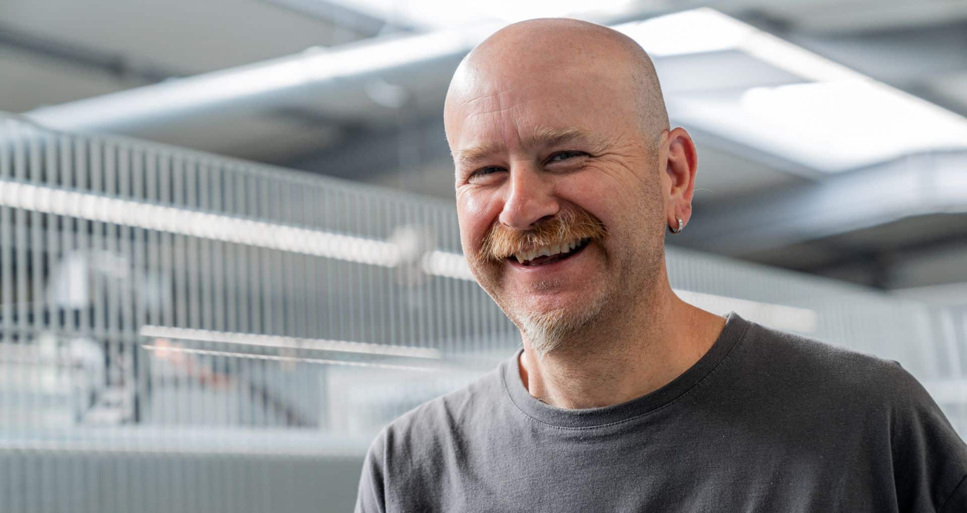 Ein lächelnder Mann mit Glatze, Schnurrbart und Ohrring steht im Haus und trägt ein graues T-Shirt.