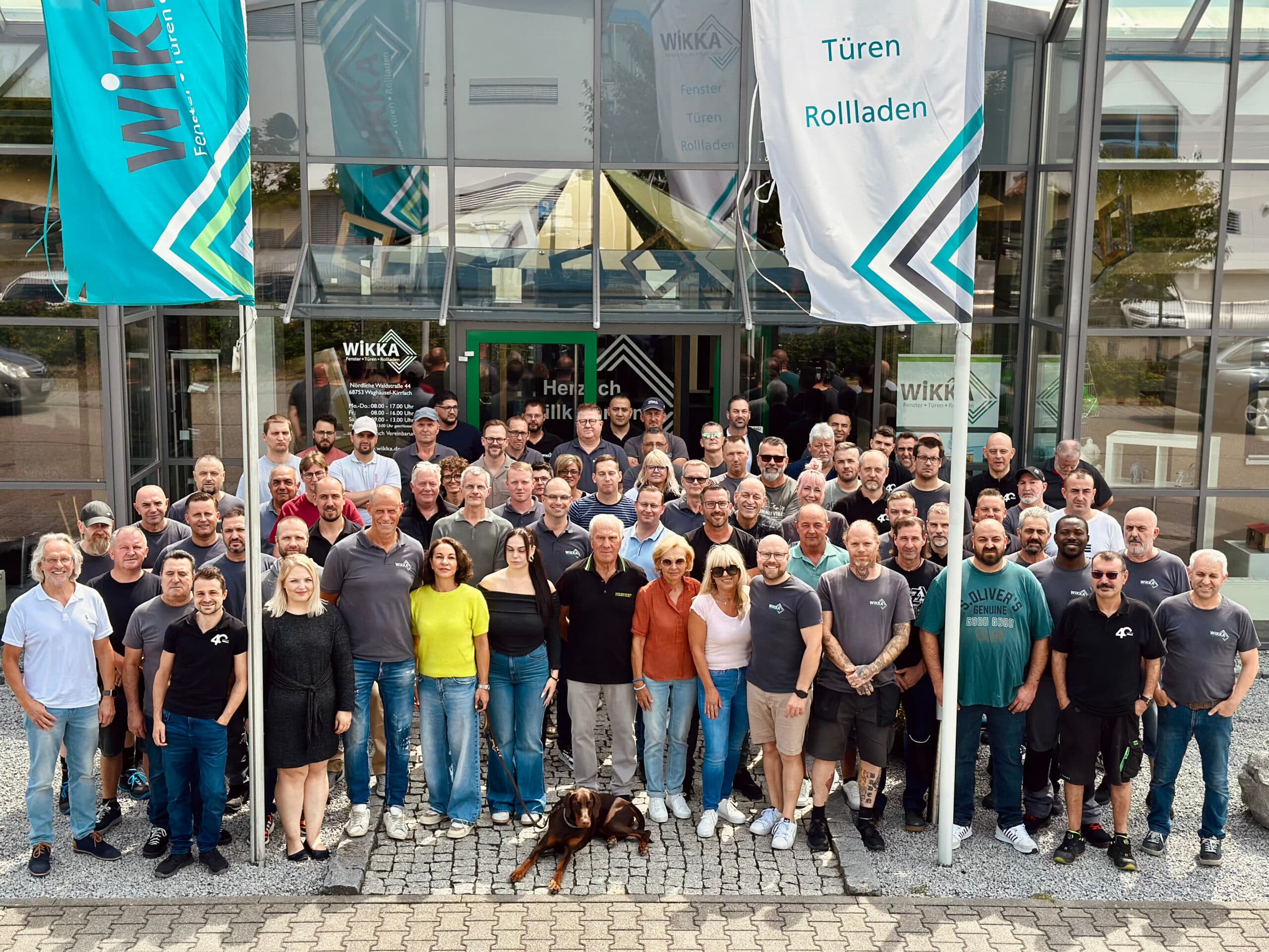 Eine gro&szlig;e Gruppe von Menschen steht l&auml;chelnd vor einem gl&auml;sernen Geb&auml;ude mit Fahnen und Schildern &uuml;ber dem Kopf.