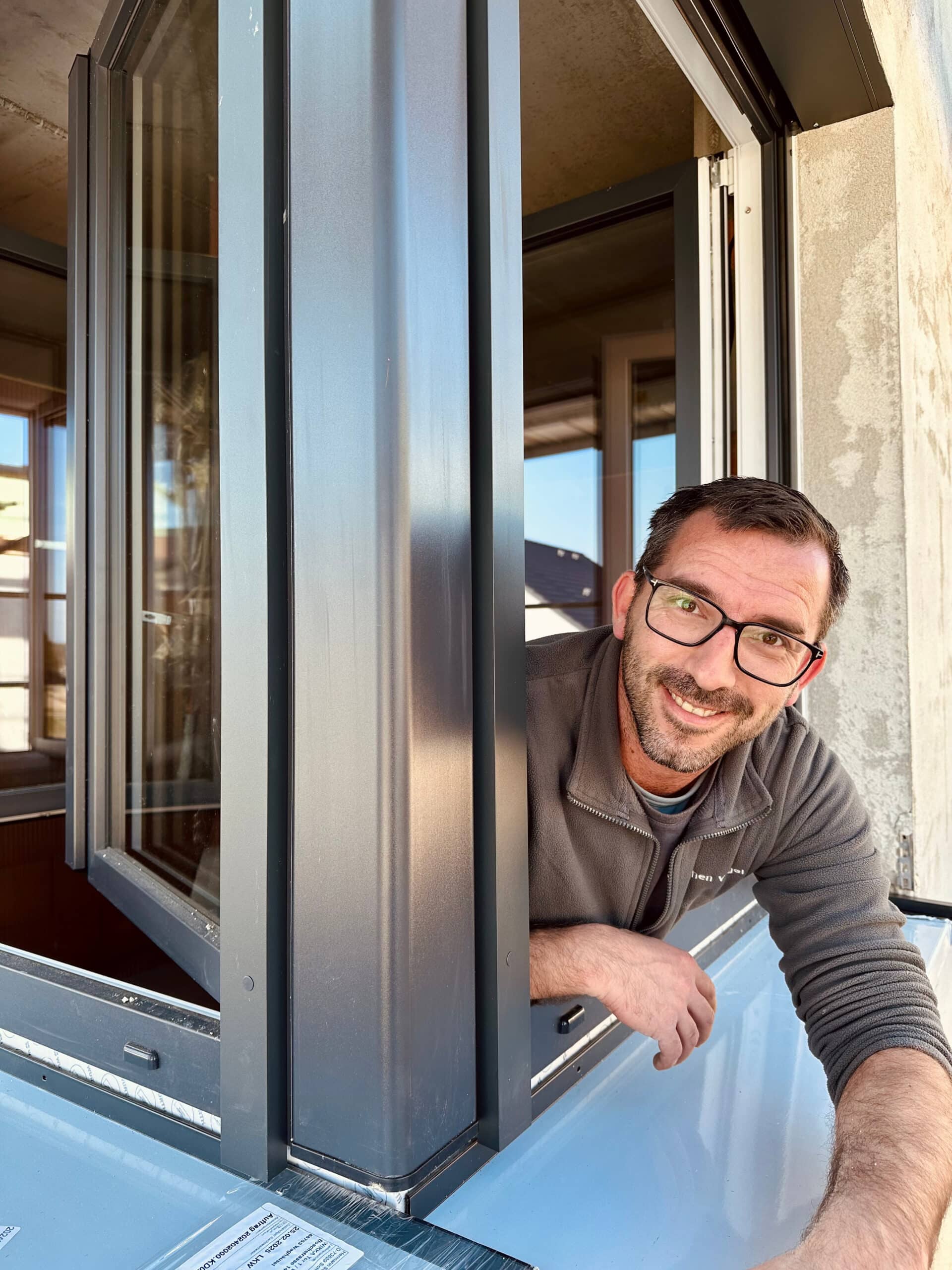 Mann mit Brille lehnt sich l&auml;chelnd aus einem offenen Fenster in einem modernen Geb&auml;ude, Sonnenlicht scheint herein.
