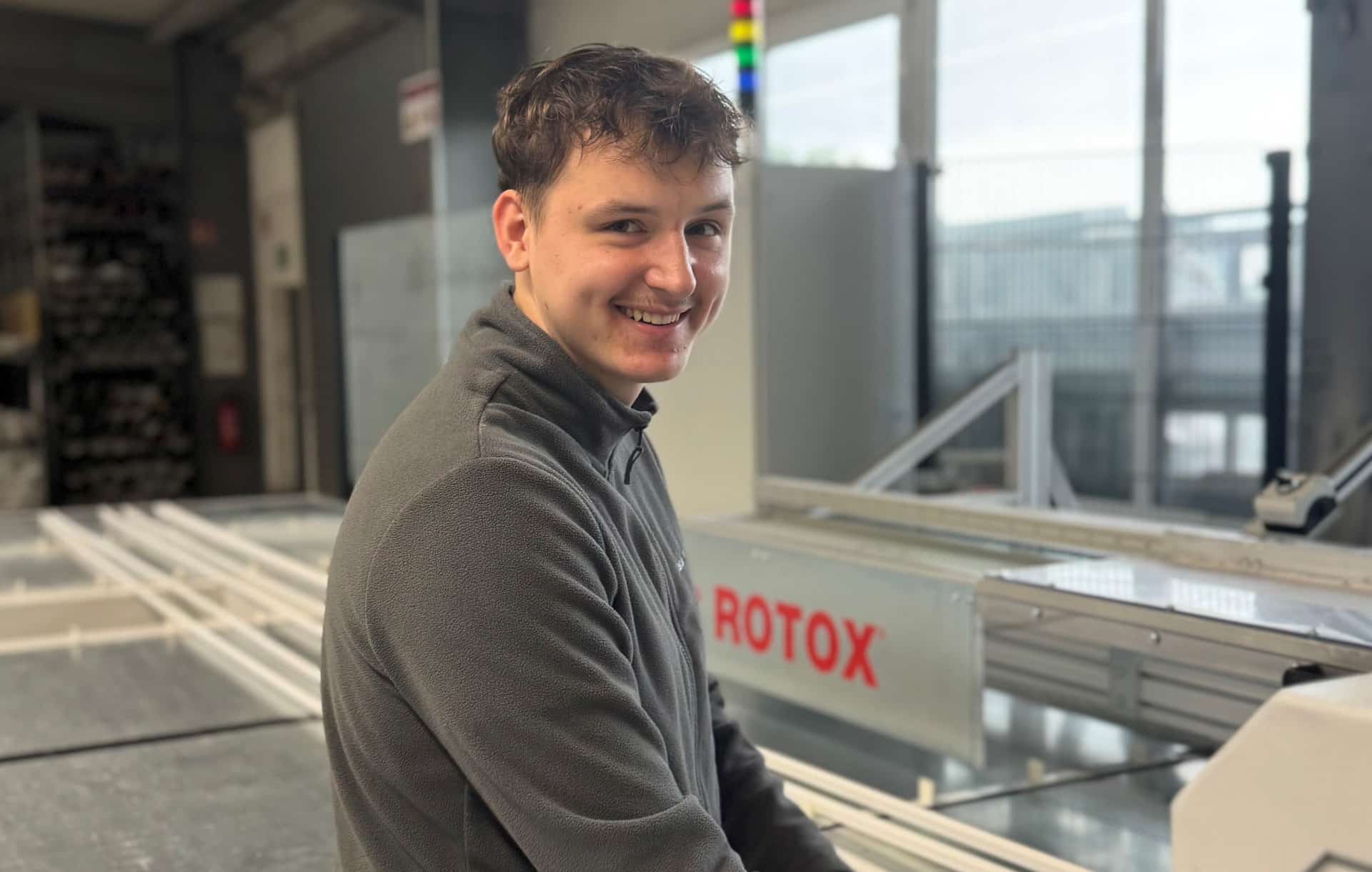 Ein junger Mann lächelt bei der Arbeit in einer Industrieanlage mit einer ROTAX-Maschine im Hintergrund.