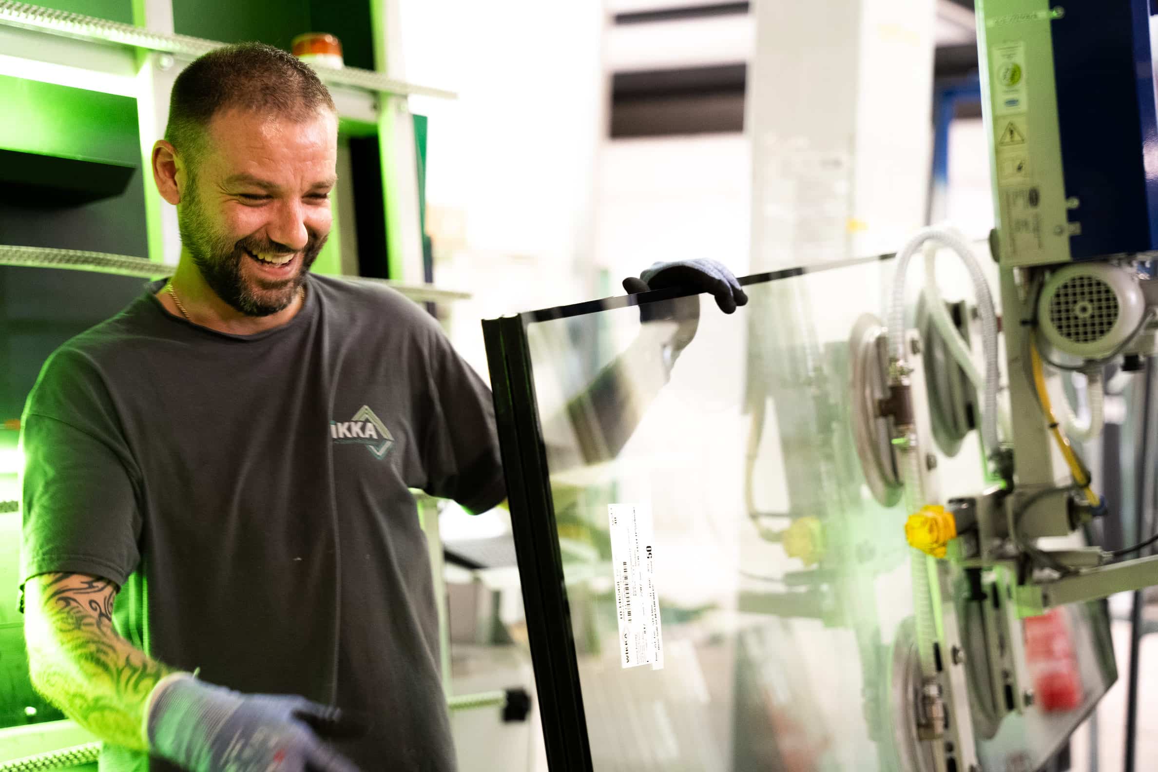 Ein l&auml;chelnder Au&szlig;endienstmitarbeiter mit Handschuhen h&auml;lt in einer hell beleuchteten Industrieumgebung eine gro&szlig;e Glasscheibe in der Hand.