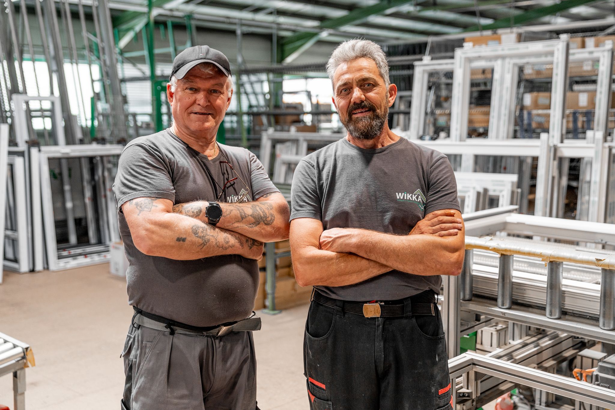 Zwei M&auml;nner in Arbeitskleidung stehen mit verschr&auml;nkten Armen in einer Fabrik voller Fensterrahmen und Maschinen und verk&ouml;rpern damit die Professionalit&auml;t, die von einem Monteur / Servicetechniker (m/w/d) in Festanstellung erwartet wird.